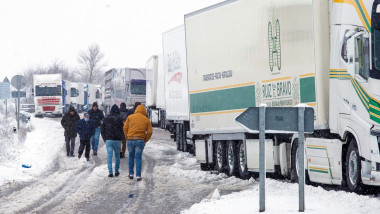 oameni care merg pe jos pe langa niste camioane care sunt parcate pe marginea unei sosele cu zapada