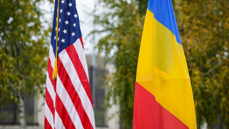 Shallow depth of field (selective focus) image with the US and Romanian flags on poles on a vegetal background.