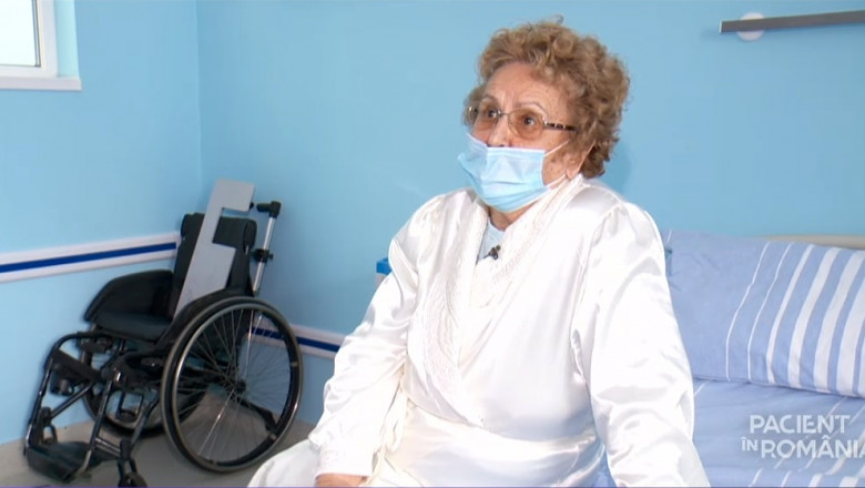 pacienta care sta pe pat in sezut intr-o camera de spital unde se vede un scaun cu rotile intr-un colt