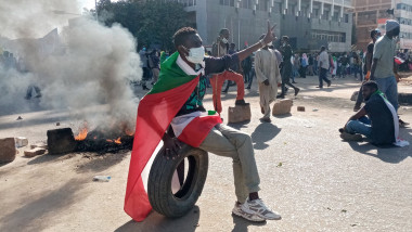 Bărbat acoperit cu steagul Sudanului stă pe un cauciuc în timp ce un foc arde în spate