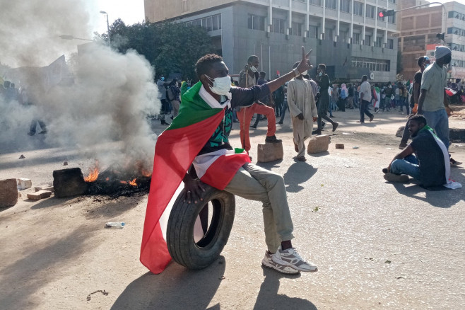 Bărbat acoperit cu steagul Sudanului stă pe un cauciuc în timp ce un foc arde în spate