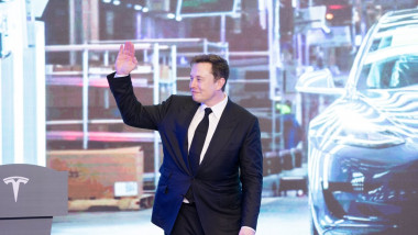 elon musk zambeste si saluta cu mana sus langa o masina tesla