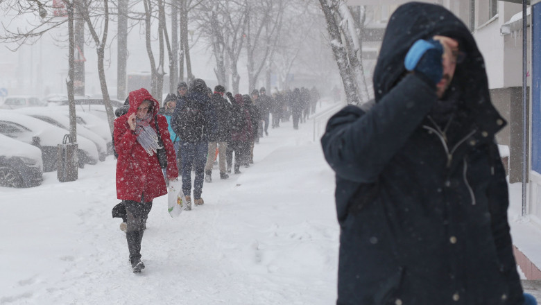 oameni merg pe trotuar acoperit de zapada in timp ce ninge
