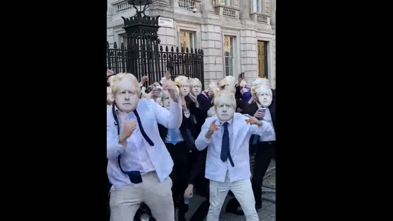 oameni cu masti cu chipul lui boris johnson dansand in fata portilor downing street