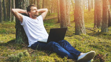 un barbat sprijinim de un copac, cu laptopul pe picioare si mainile la ceafă, relaxat