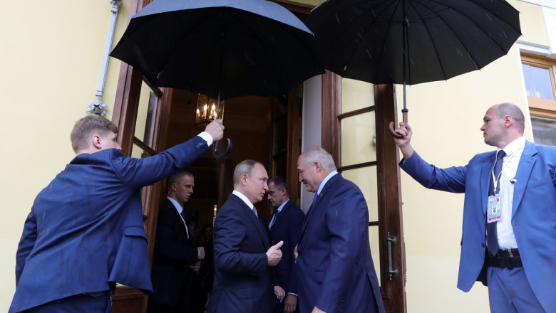 Aleksandr Lukașenko și Vladimir Putin dau mana, in prezenta unor membri ai delegatiilor care le tin umbrelele