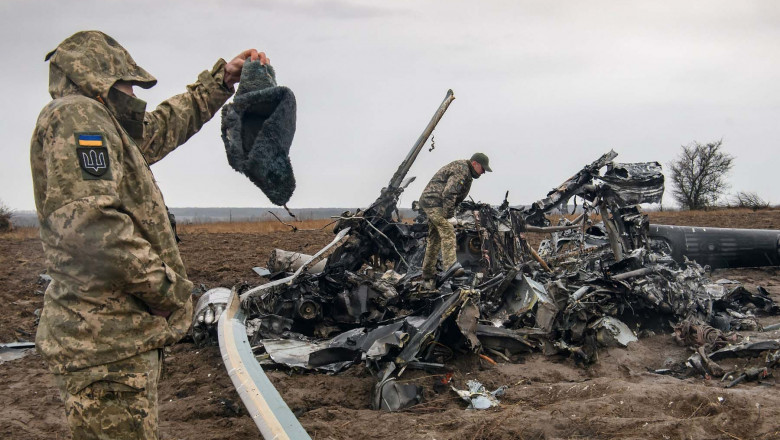 2 soldati ucraineni cerceteaza carcasa unui elicopter rus doborat pe camp. Unul din ei tine in mana o caciula ruseasca de militar