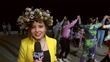 reporter cu o coroana de flori pe cap in spatele caruia danseaza oameni prinsi intr-o hora