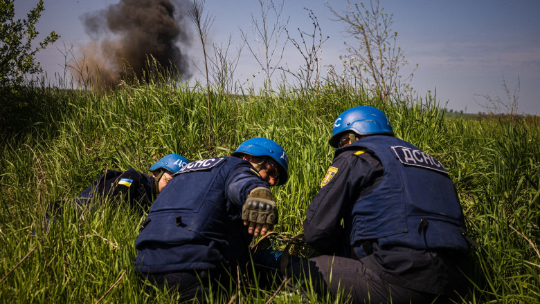 bărbați din forțele speciale îngenunchează în zona unei mine neexplodate în timp ce se vede o explozie cu fum negru pe fundal