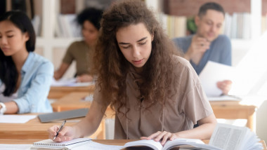 Teen girl studying with textbook writing essay learning in classroom