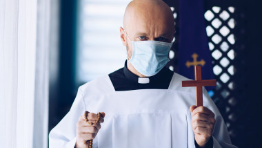 Man priest in medical mask praying with cross and rosary.