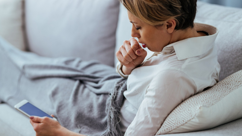 femeie bolnava care sta in pat, tuseste si tine un smartphone in mana