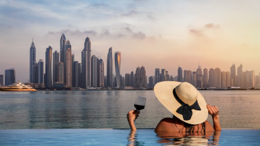 A elegant woman with a hat and a drink in her hand stands at the edge of a infinity pool and enjoys the view to the skyline of the Dubai Marina during sunset, UAE