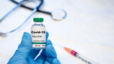 Fotografie ilustrativă cu un medic care ține în mâna acoperită de mânușă sanitară o doză de vaccin anti-coronavirus. Pe fundal sunt vizibile o seringă și un stetoscop.