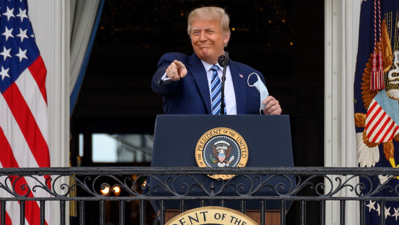 Președintele Donald Trump zâmbește de la balconul Casei Albe în perioada îmbolnăvirii sale cu Covid-19, ținând masca în mână și arătând cu degetul.