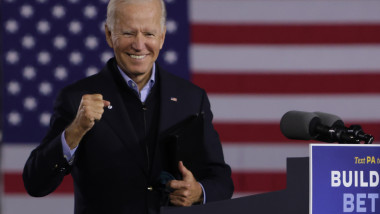 Joe Biden zâmbește la tribună, în cadrul unui eveniment de campanie, cu drapelul Statelor Unite ale Americii pe fundal.