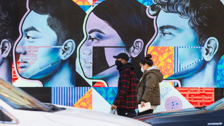 oameni pe strada cu masti pe fata merg in fata unui zid pictat cu figuri ale oamenilor care poarte masca