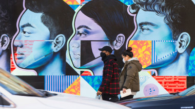 oameni pe strada cu masti pe fata merg in fata unui zid pictat cu figuri ale oamenilor care poarte masca