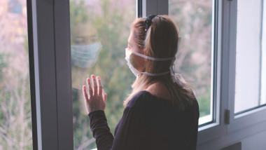 Femeie cu mască de protecție privește pe geam în timpul unui lockdown anti-coronavirus