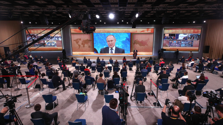 o multime de jurnalisti din rusia, intr-o sala in care se uta la un ecran urias pe care este proiectat vladimir putin care le raspunde la intrebari prin videocoferinta