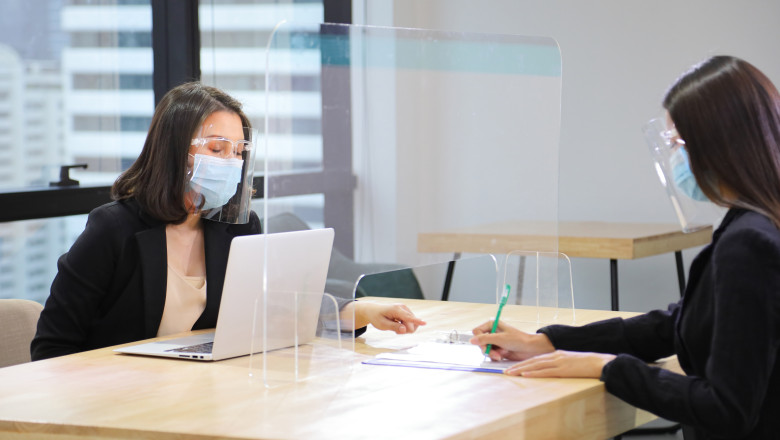 Manager from HR department wearing facial mask is interviewing new applicant who is handing her resume and profile through the partition for social distancing and new normal