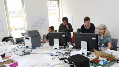 Mai mulți angajati la un birou stau in fața unor calculatoare