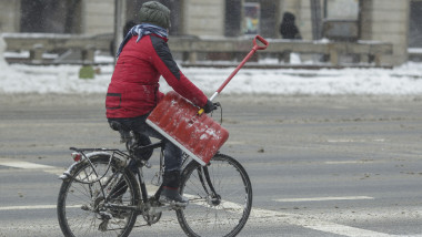 un barbat merge cu o lopata in mana pe bicicleta, pe timp de ninsoare.