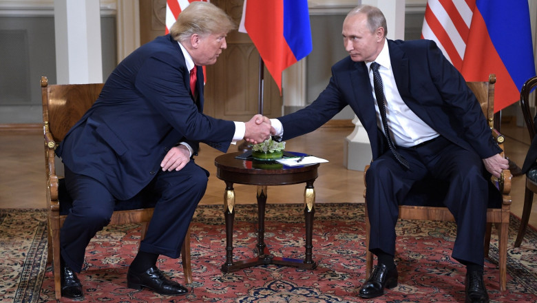 Cadru oficial. În sală, în fundal, se văd steagurile Statelor Unite și al Federației Ruse. În primplan, stând pe scaune, Donald Trump și Vladimir Putin își strâng mâna.