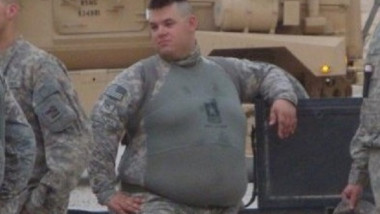 Pe fundal, un transportor blindat în curtea unei baze militare americane. Un soldat american gras se sprijină cu cotul de portiera unei mașini.