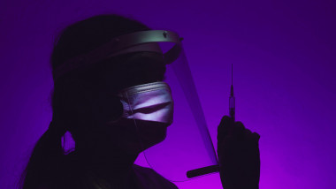 silueta unei doctorite cu masca tinand in mana o seringa, pe un fond mov intunecat