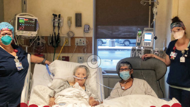 Doi vârstnici pe paturile de spital, tinandu-se de mână, alături de două asistente ale spitalului în care cei doi sunt internati cu covid