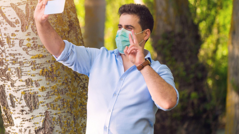 Bărbat cu mască de protecție își facă un selfie gesticulând semnul victoriei într-un parc