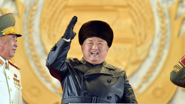 Kim Jong Un, îmbrăcat gros și purtând o căciulă rusească, salută militarii. Pe fundal se vede emblema de stat a Coreei de Nord.