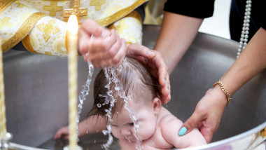 un preot toarna apa cu palmele in capul unui copil aflat in cristelnita de botez