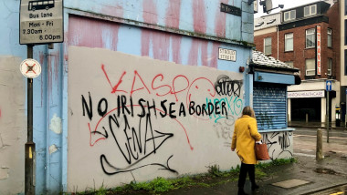 femeie cu jacheta galbena trece pe langa un zid cu graffiti no irish sea border in centrul belfastului
