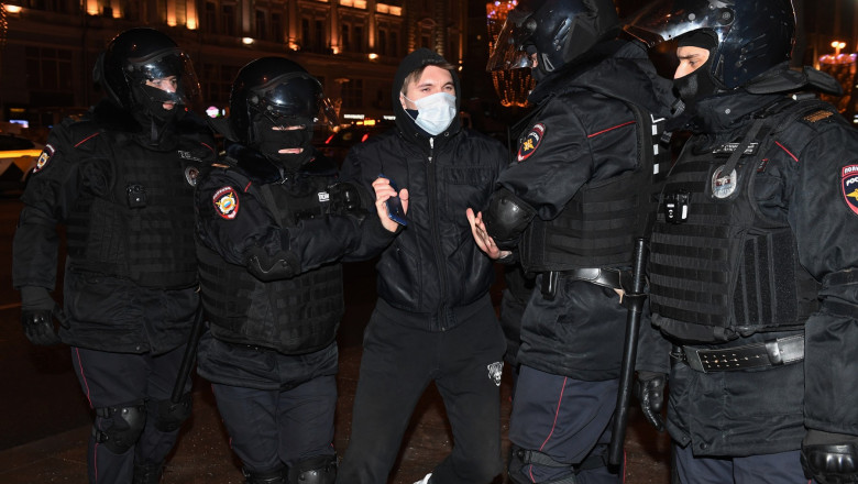 Un bărbat cu mască de protecție și îmbrăcat în negru este ținut de mâini de doi polițiști ruși îmbrăcați în echipament special.