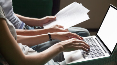un tanar scrie la un laptop pe picioare iar altul sta langa cu documente in maini