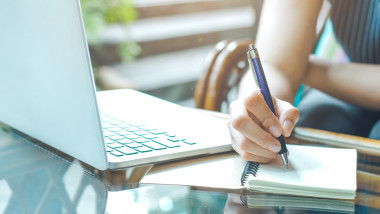 mana de femeie langa un laptop, care notează ceva intr-un caiet scriind de mana