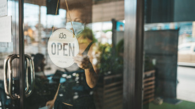 angajata cu masca la usa unui restaurant avand mana pe cartonul pe care scrie „open”