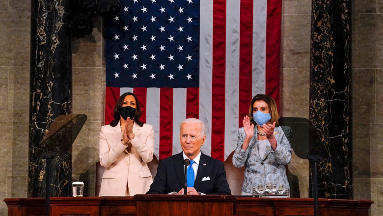 Joe Biden pe podium, ]n spate sunt vicepreședinta Kamala Harris și lidera Camerei Reprezentanților Nancy Pelosi, cu steagul SUA pe perete