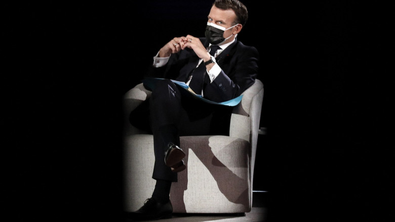 Emmanuel Macron, stă pe un fotoliu alb, cu mască de protecție neagră, și mâinile împreunate