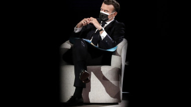 Emmanuel Macron, stă pe un fotoliu alb, cu mască de protecție neagră, și mâinile împreunate