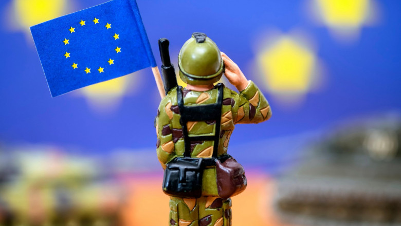 O figurină soldățel cu steagul UE în mână. Pe fundal se văd figurine de tanc