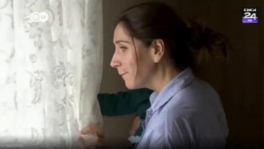 femeie cu copil in brate care se uita pe fereastra de dupa o perdea