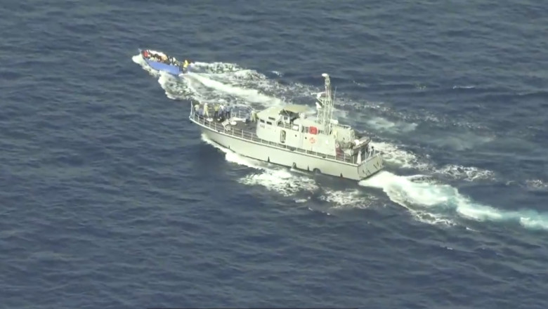 o barcă albastră din lemn cu un motor mic și cel puțin 20 de oameni la bord este urmărită în cercuri, cu viteză mare, de către Paza de coastă libiană.