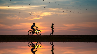 un barbat pe bicicleta merge in spatele unei fete care alearga pe marginea apei la apus