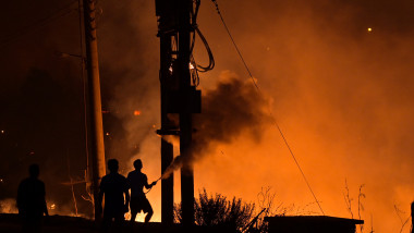 Pompieri care se văd ca niște siluete într-o atmosferă portocalie, încearcă să stingă incendiul masiv din apropierea atenei din august 2021