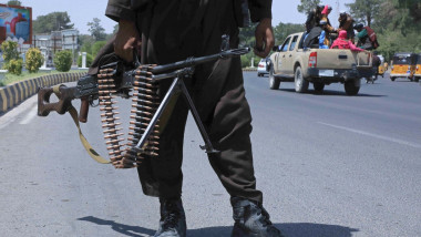 luptator taliban cu o arma in mana pe o sosea in timp ce in fundal se vede o masina pickup cu afgani in spate
