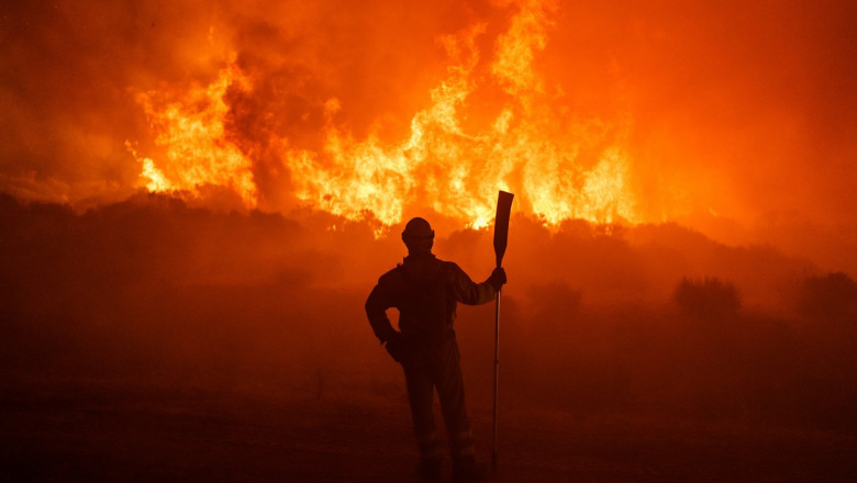 pompier cu loptata priveste la flacarile unui incendiu de vegetație