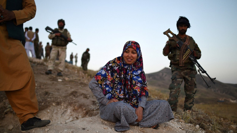 Femeie cu capul acoperit stă turcește pe marginea unei stânci. În spate e un bărbat echipat cu mitralieră și muniție
